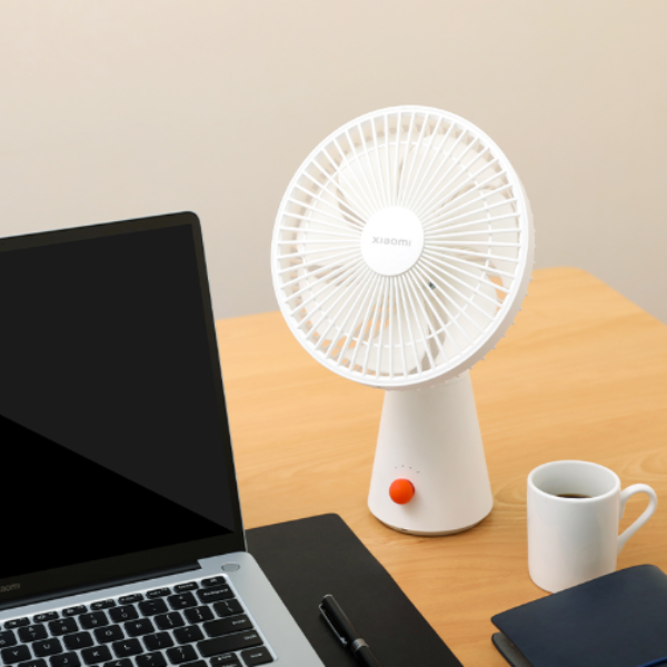 White portable fan on a desk with a laptop, mug, and notebook.