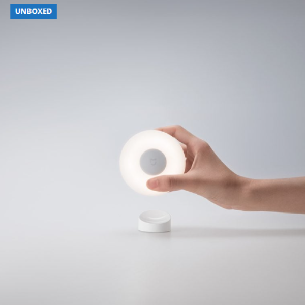 Hand holding a white round device with a light on a gray background, branded 'UNBOXED'.