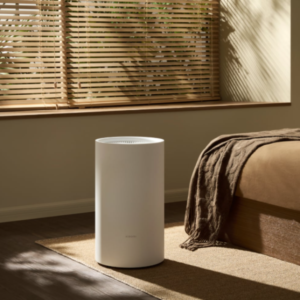 White cylindrical air purifier on a wooden floor with a couch and window blinds in the background