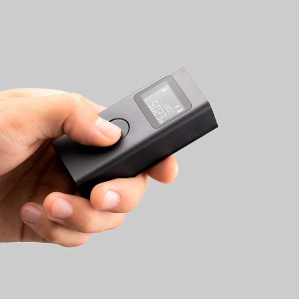 Hand holding a black electronic device with a digital display on a gray background