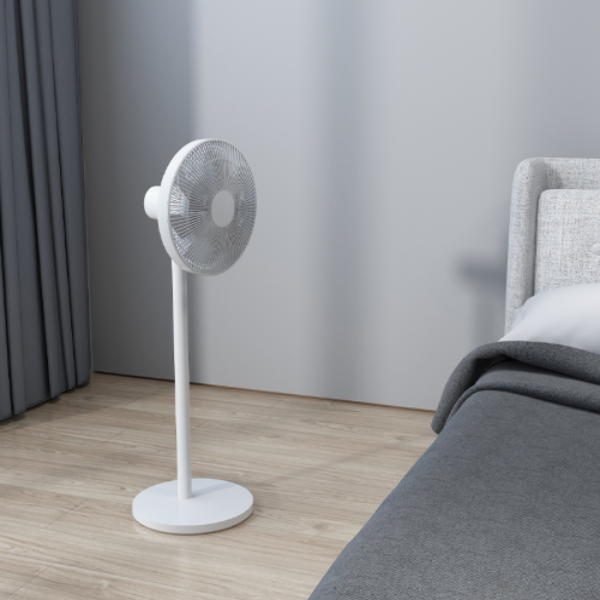 White standing fan in a bedroom setting with a bed and curtains.
