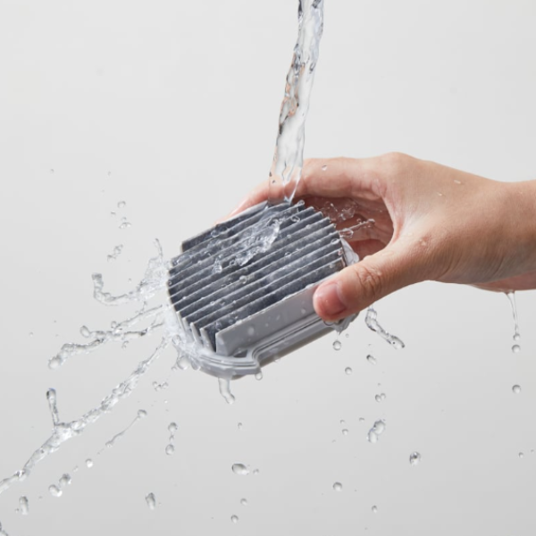 Hand holding a metallic heat sink under running water on a light gray background
